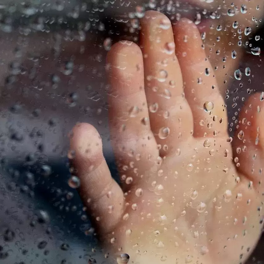 A child's hand resting on a window with water drops on the opposite side of the glass.